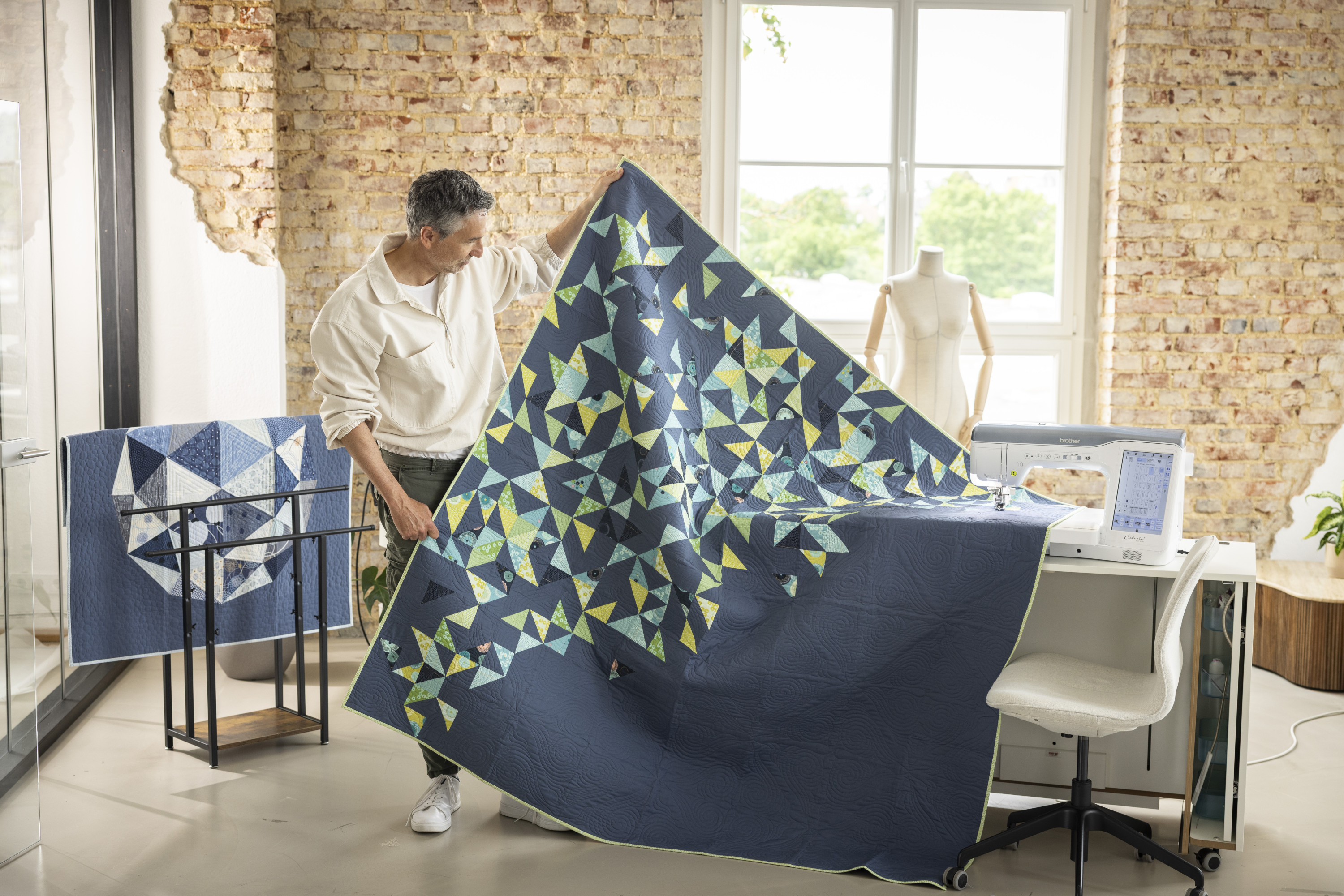 Man holding a large blue and green patterned fabric in a room with a sewing machine and mannequin.