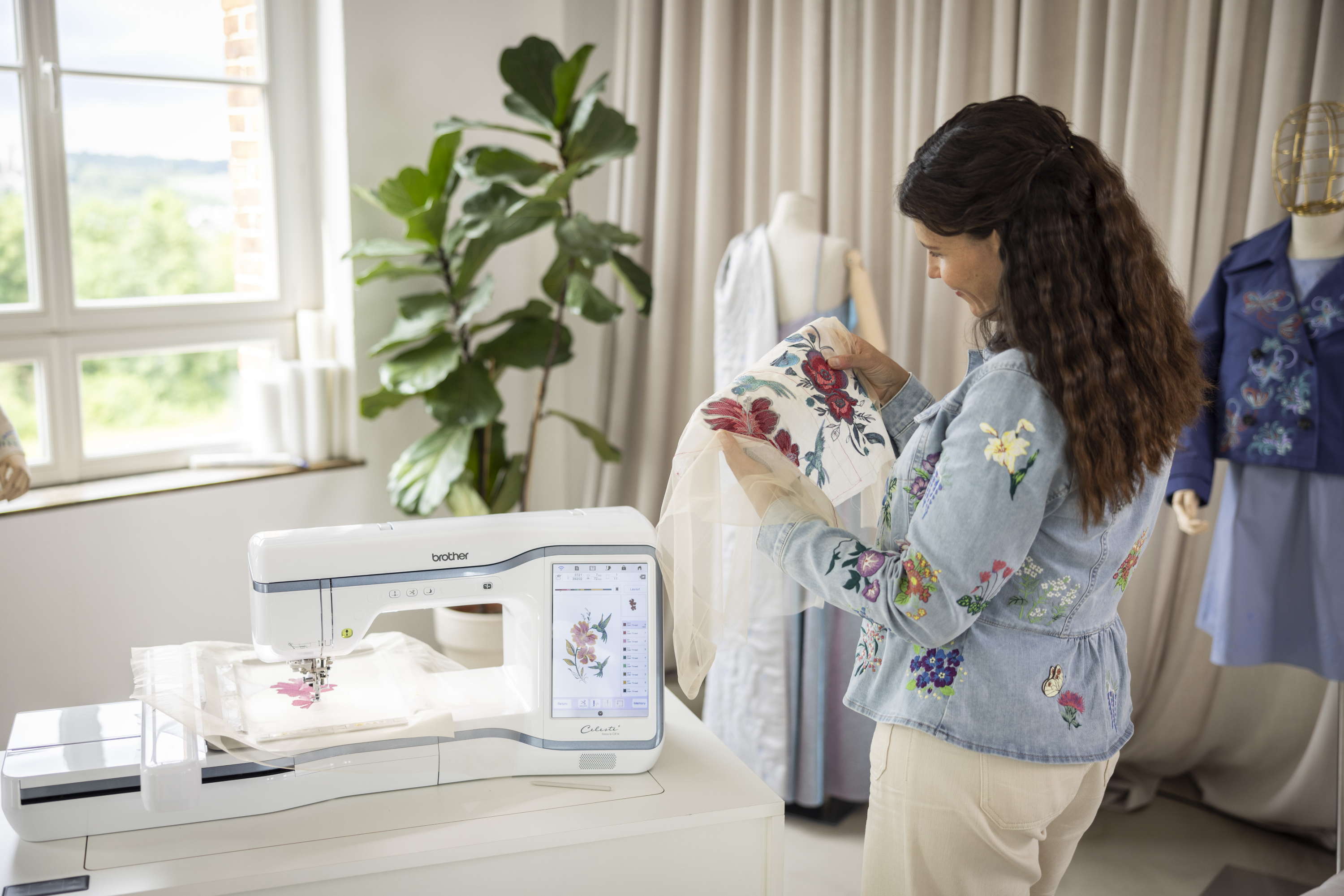 Woman working with embroidery machine and fabric in a bright room with plants and mannequins.