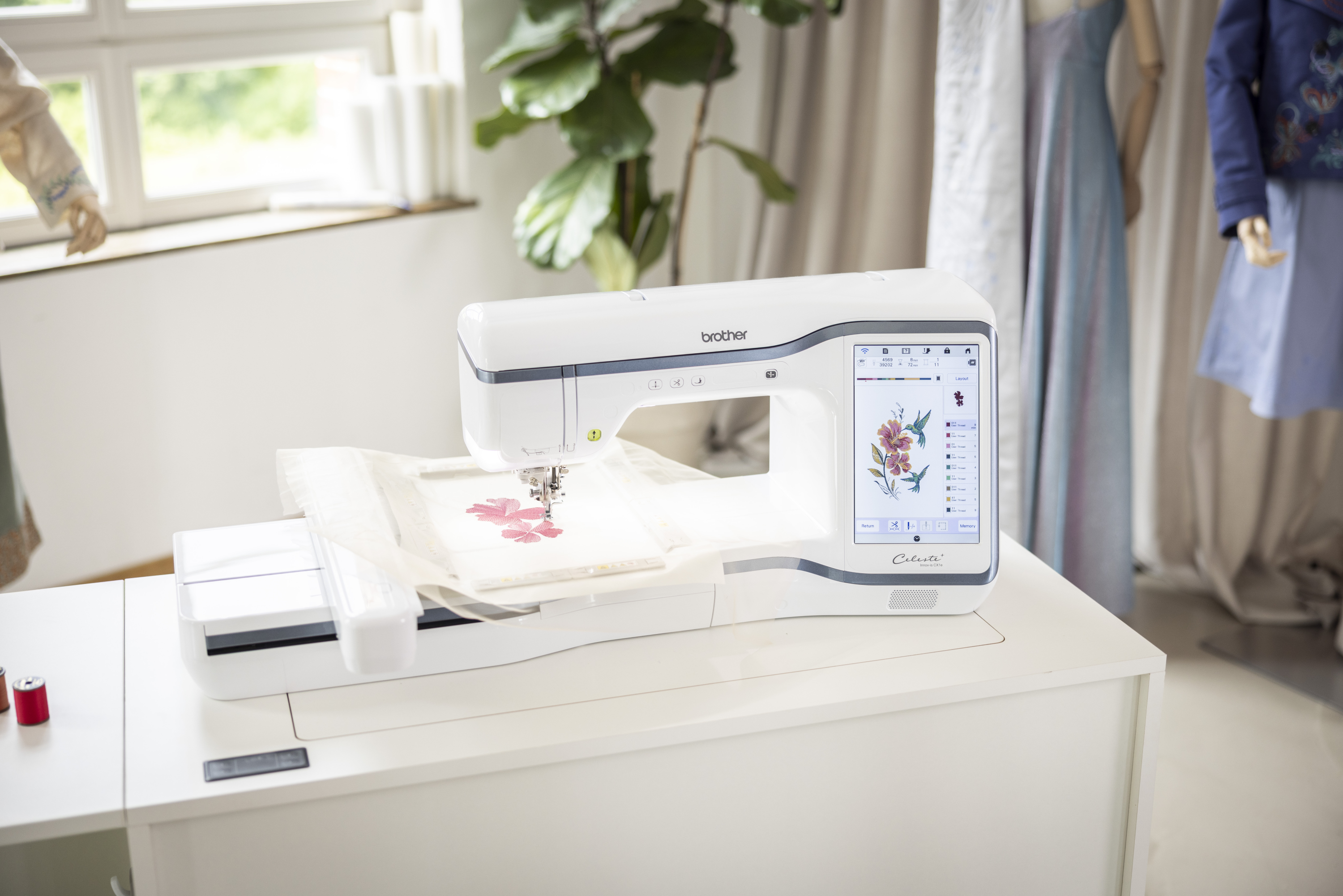 Brother embroidery machine on a white table with a plant and window in the background