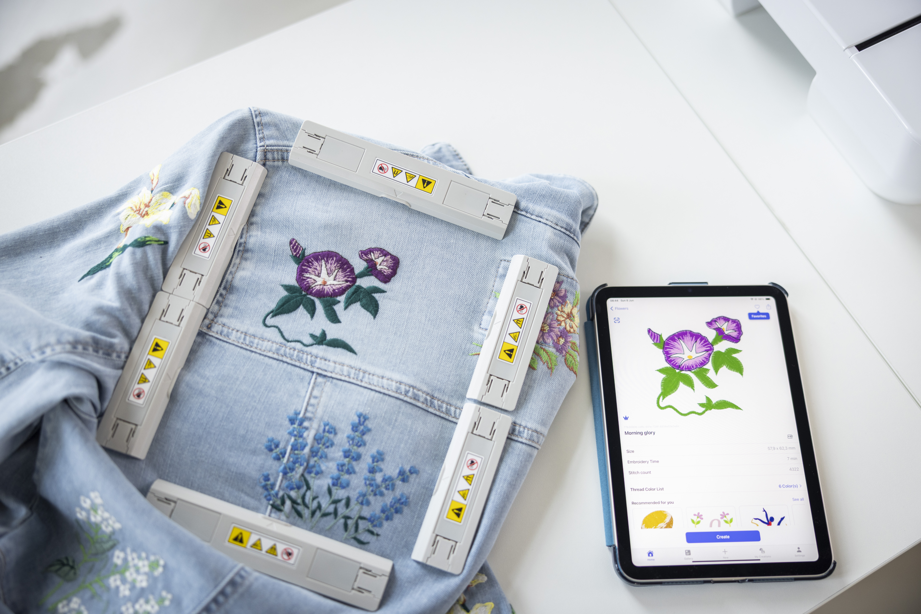 Embroidered denim fabric with floral designs next to a tablet displaying embroidery software on a white surface.