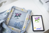 Embroidered denim fabric with floral designs next to a tablet displaying embroidery software on a white surface.
