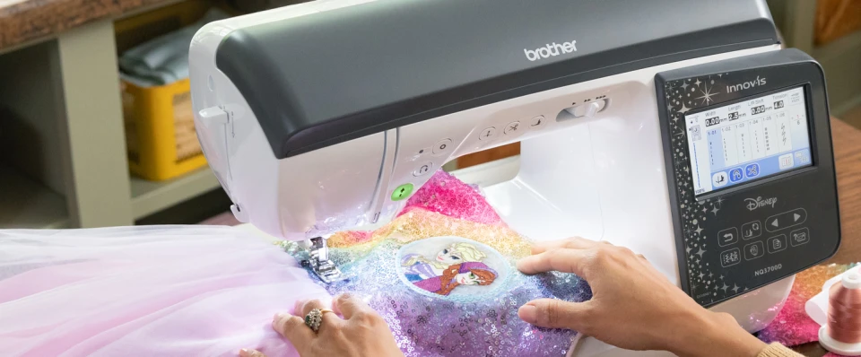 A Brother Innov-is sewing machine in use, stitching a colorful glittery fabric with a person's hands guiding it. The machine has a white and dark gray body with a digital control screen on the right.