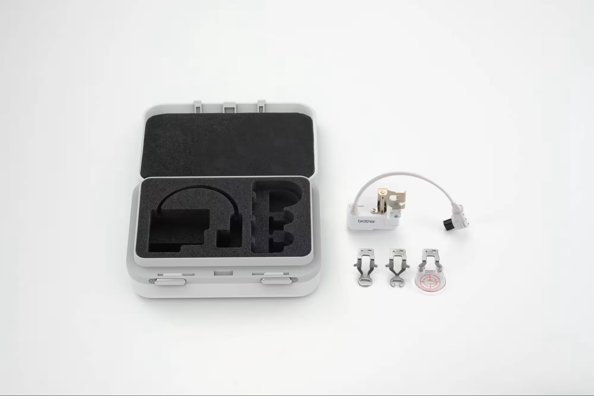 An open gray hard carrying case with black foam inserts, shown next to a small white and gray stitch regulator, three metal sewing feet, and a small round accessory on a clean white background.