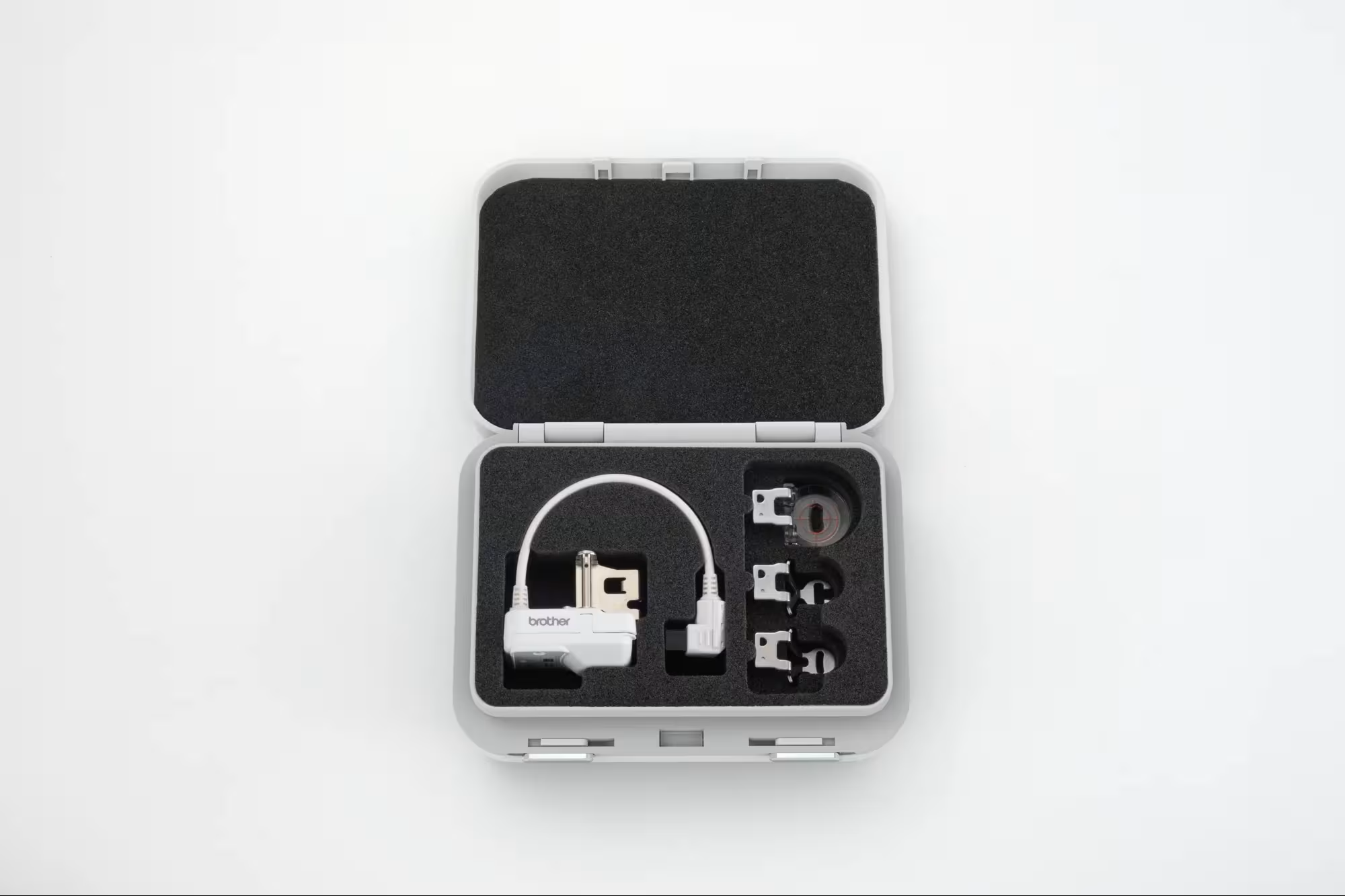 An open gray hard carrying case with black foam insert, containing a small white stitch regulator and several small sewing feet, photographed on a clean white background.