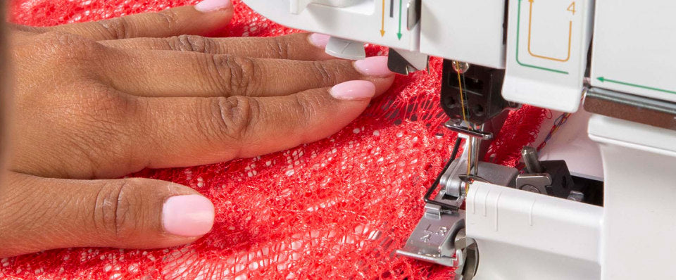 A close-up of a person's hand with light pink manicured nails guiding a red textured fabric under the presser foot of a white sewing machine.