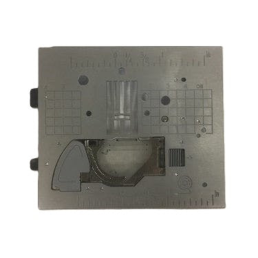 A square gray metal needle plate with various engraved markings, grid patterns, and a central transparent bobbin area, isolated on a white background.