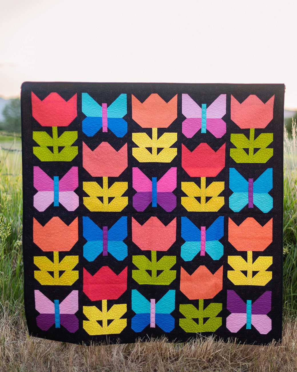 A square quilted patchwork blanket or small quilt with a black background and rows of tulips and butterflies in bright colors, photographed outdoors on dry grass with a blurred field and sky behind it.