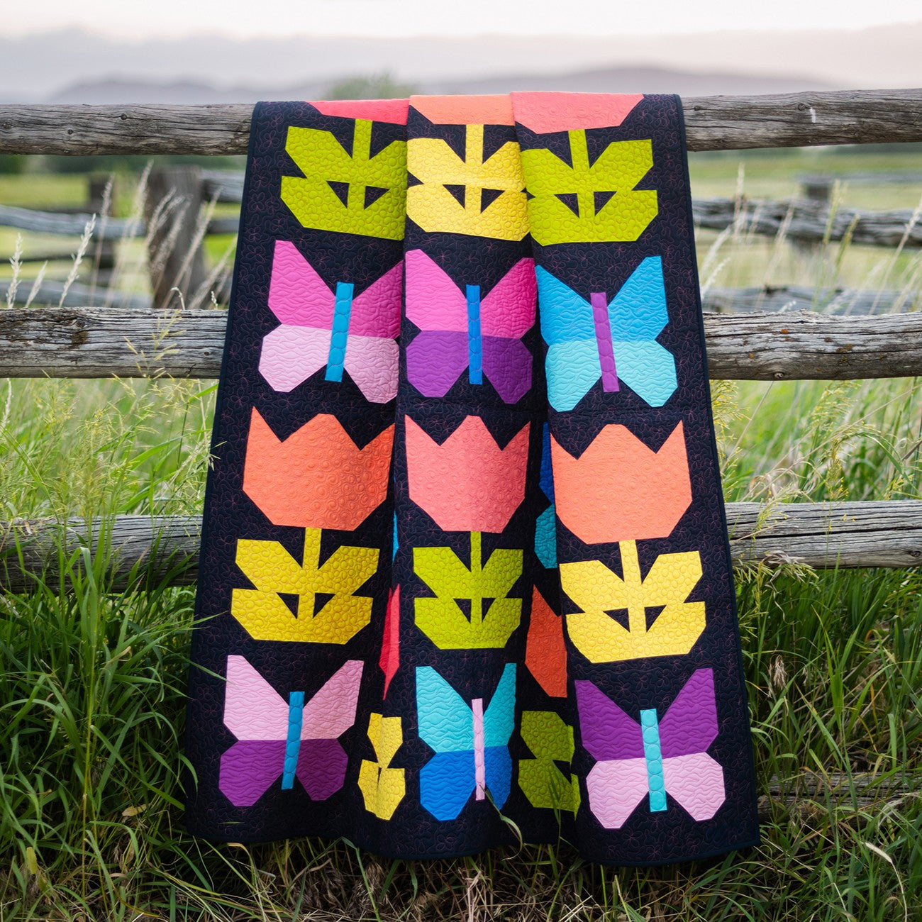 A colorful quilted blanket with tulips and butterflies draped over a rustic wooden fence in a grassy field.