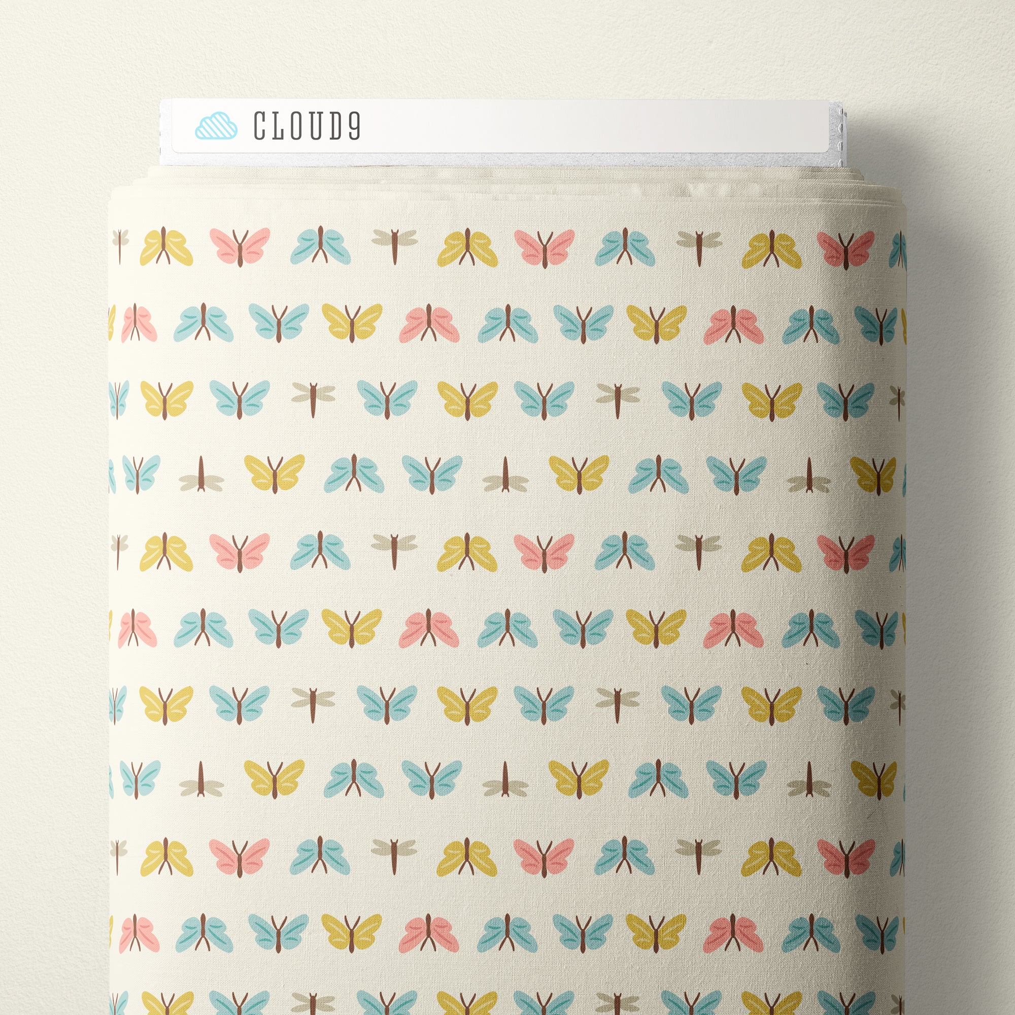 A roll of Cloud9 fabric with a repeating pattern of colorful dragonflies on a warm cream background, shown against a neutral light beige wall.