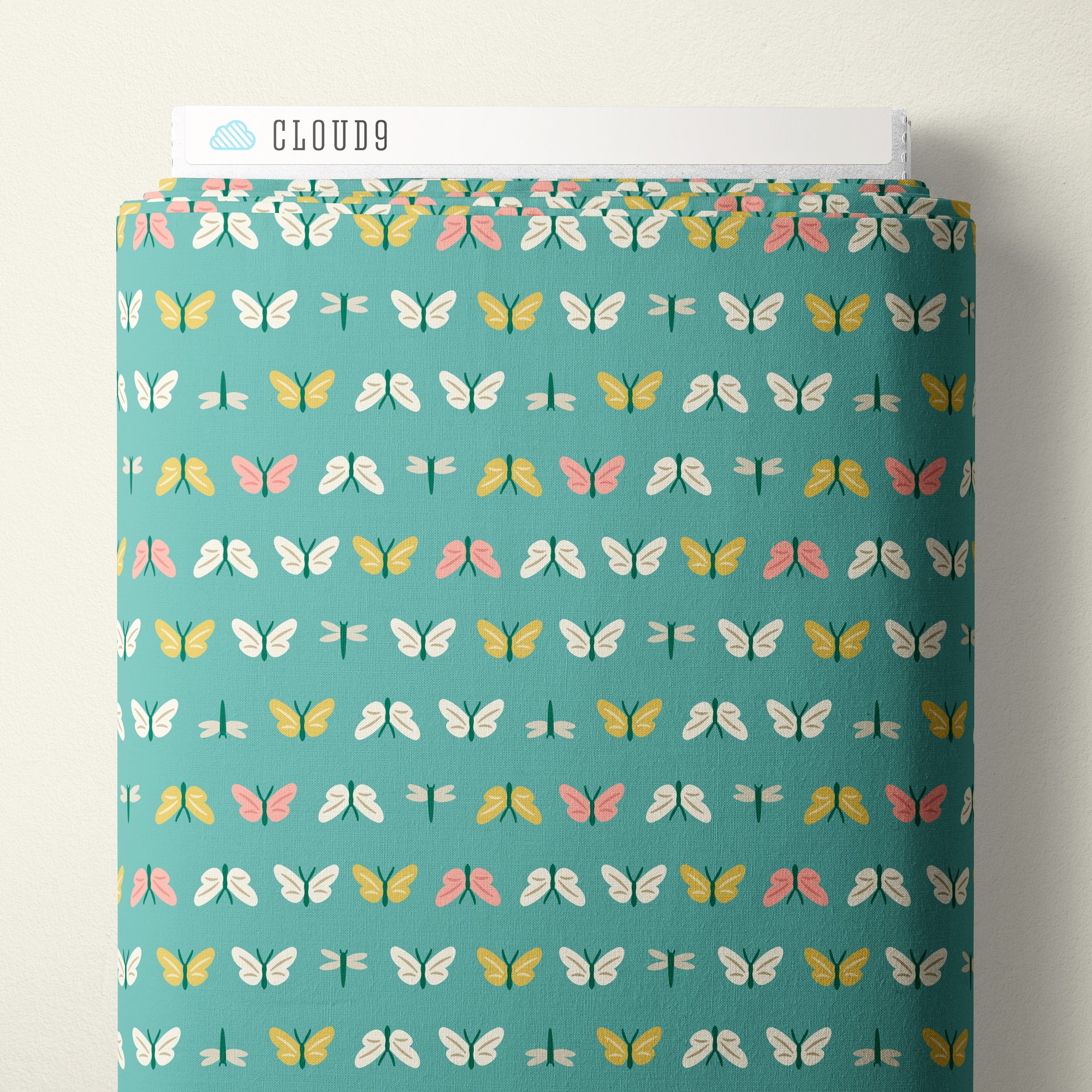 A roll of fabric with a repeating pattern of butterflies and dragonflies on a teal background, with a white header card labeled CLOUD9 at the top, photographed against a plain light wall.