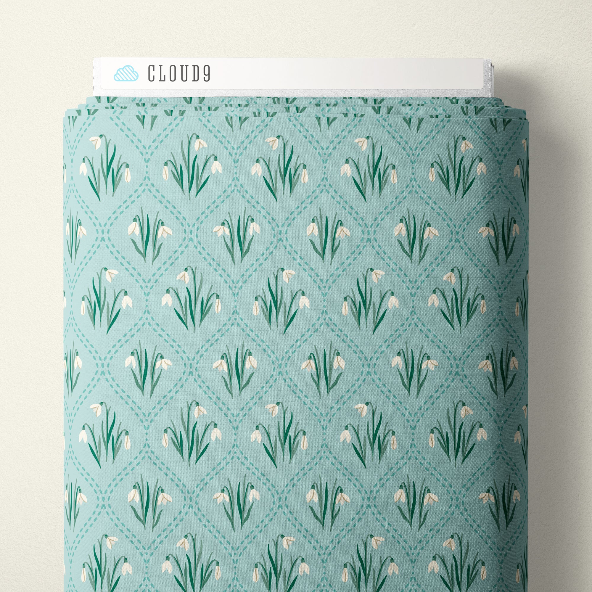 A roll of Cloud9 fabric with a repeating snowdrop pattern on a light teal background, shown against a neutral off-white wall.