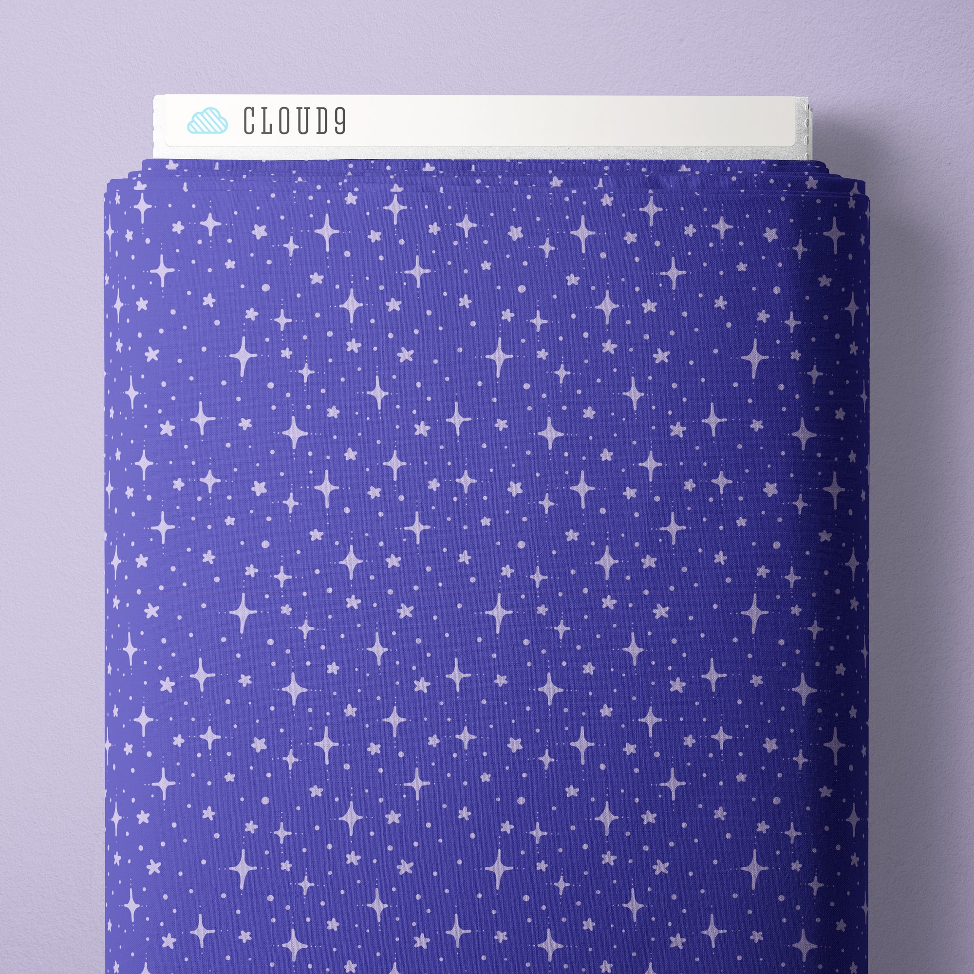 A close-up of a fabric swatch with a repeating white star and starburst pattern on a periwinkle blue background, shown against a light lavender wall. A white label at the top reads CLOUD9.