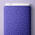 A close-up of a fabric swatch with a repeating white star and starburst pattern on a periwinkle blue background, shown against a light lavender wall. A white label at the top reads CLOUD9.