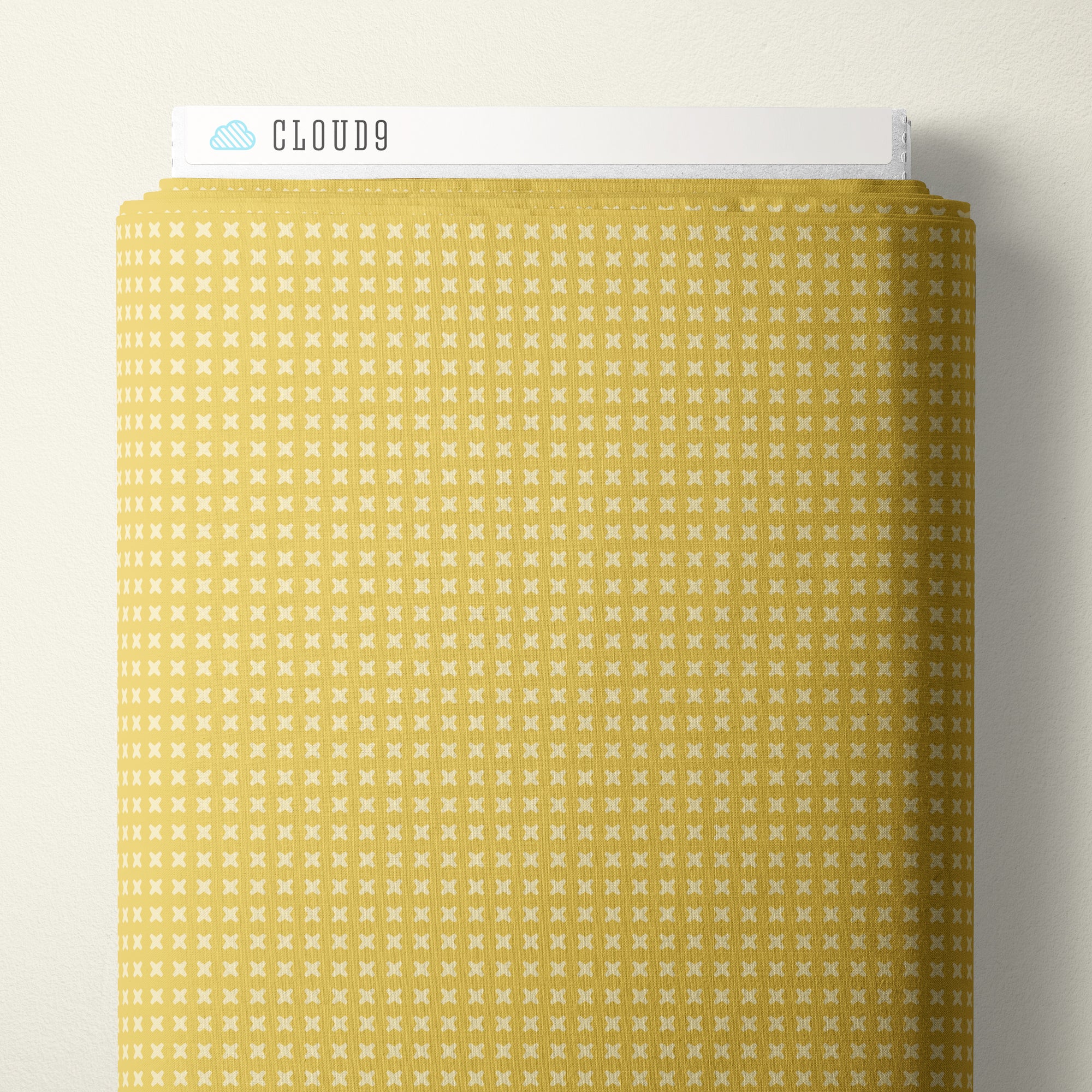 A roll of patterned fabric with a repeating small white x motif on a warm yellow background, shown against a light neutral wall. A white header card at the top reads CLOUD9.