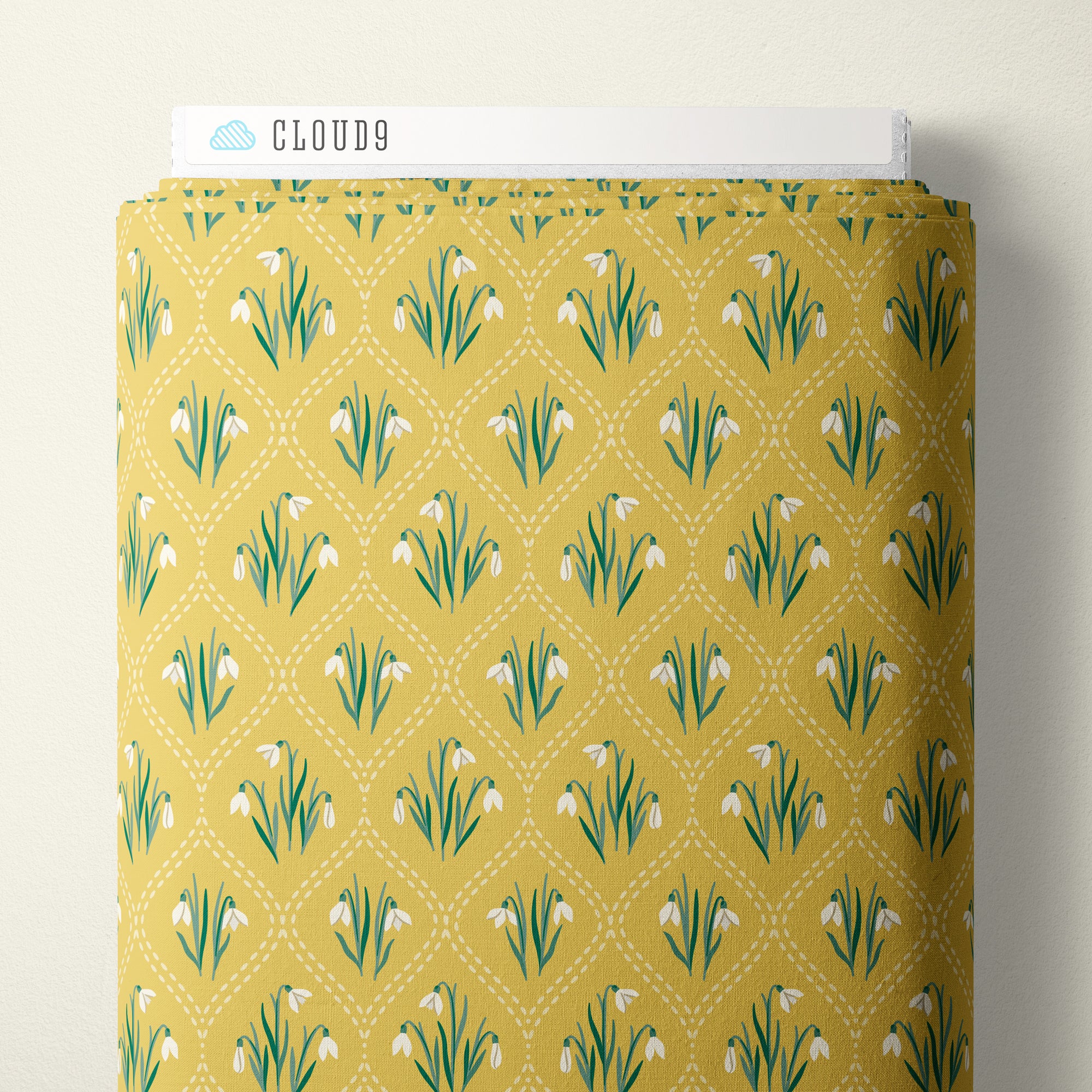 A roll of fabric with a repeating pattern of white snowdrops on a mustard yellow background, featuring green stems and leaves and a white dotted diamond lattice. A white label at the top of the roll is printed with the brand name 'CLOUD9'.
