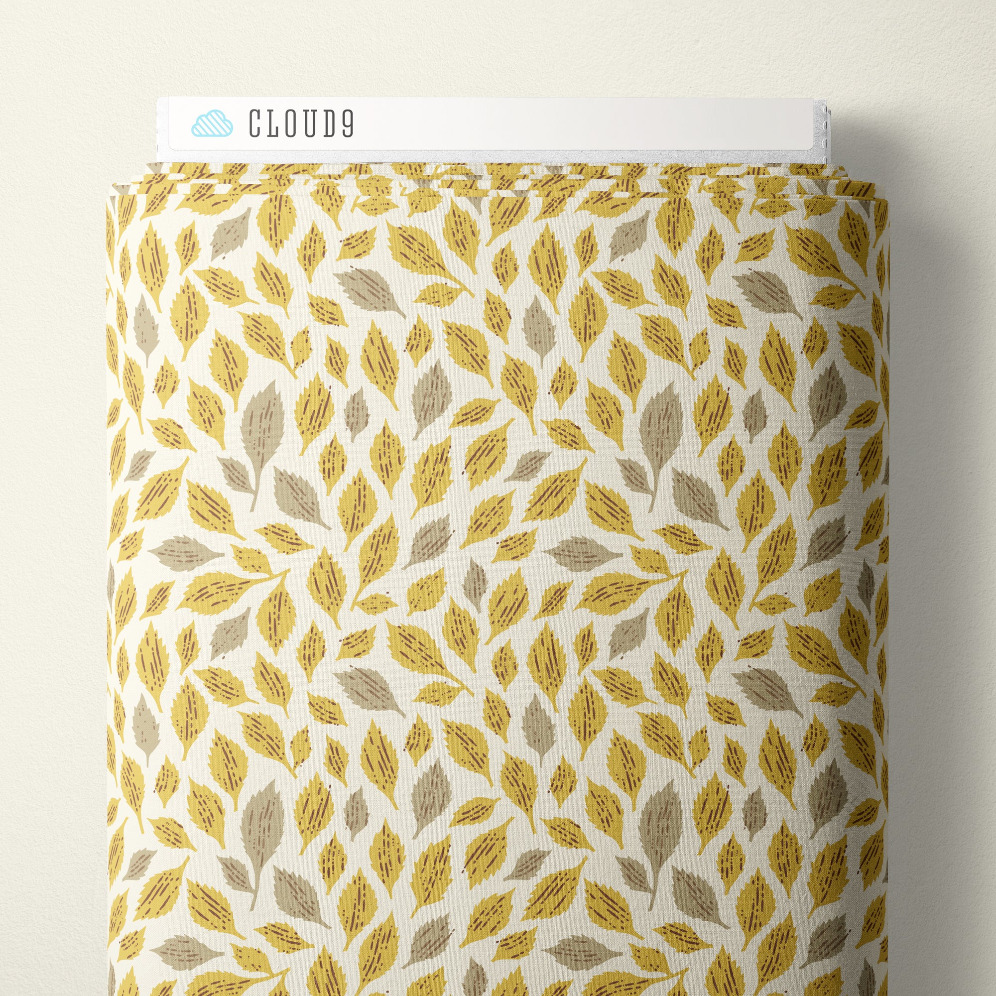 A roll of Cloud9 fabric featuring a repeating pattern of mustard yellow and taupe leaves on a warm off-white background, shown against a neutral beige wall.