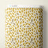 A roll of Cloud9 fabric featuring a repeating pattern of mustard yellow and taupe leaves on a warm off-white background, shown against a neutral beige wall.