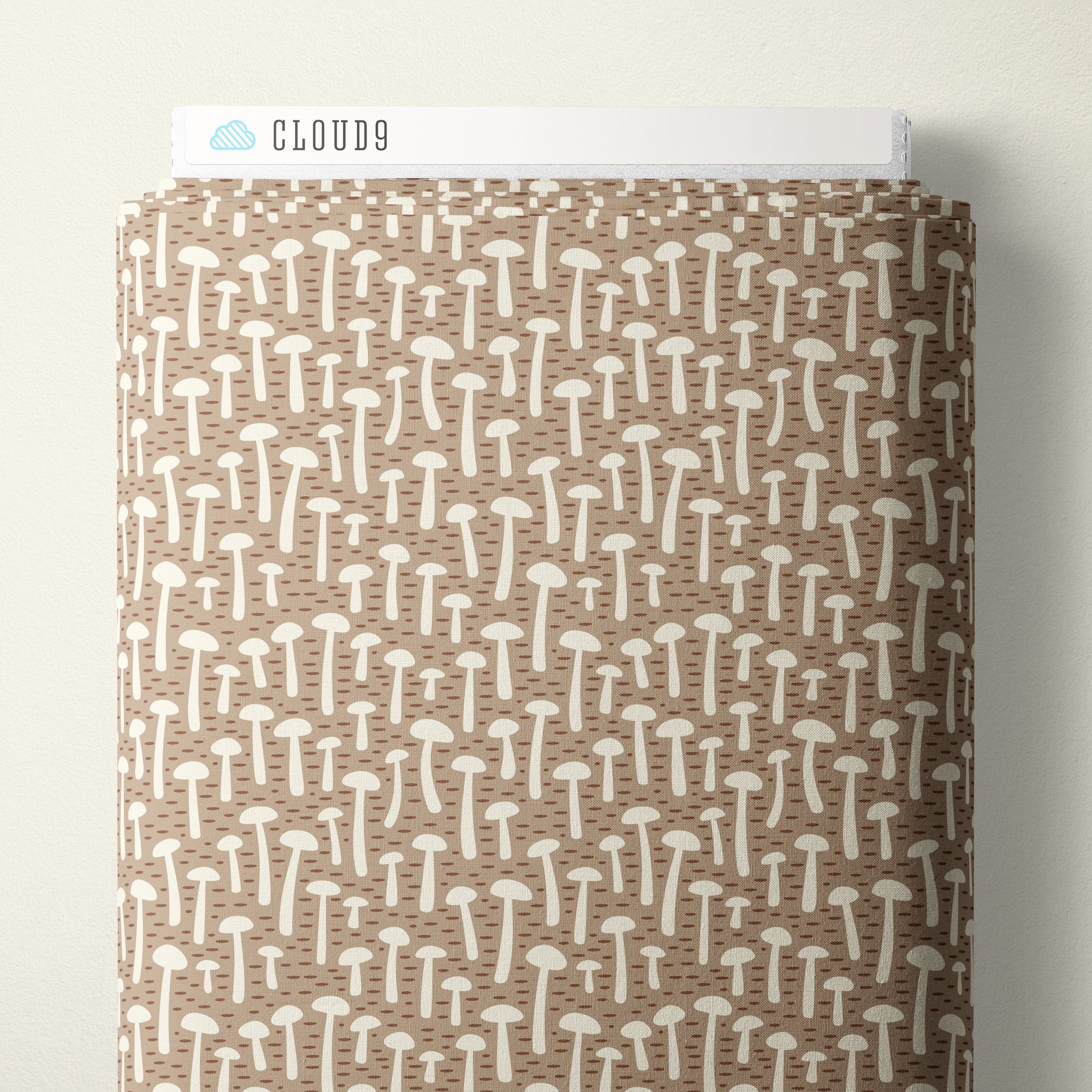 A roll of Cloud9 brand fabric featuring a repeating pattern of small white mushrooms on a warm brown background, shown against a neutral off-white wall.