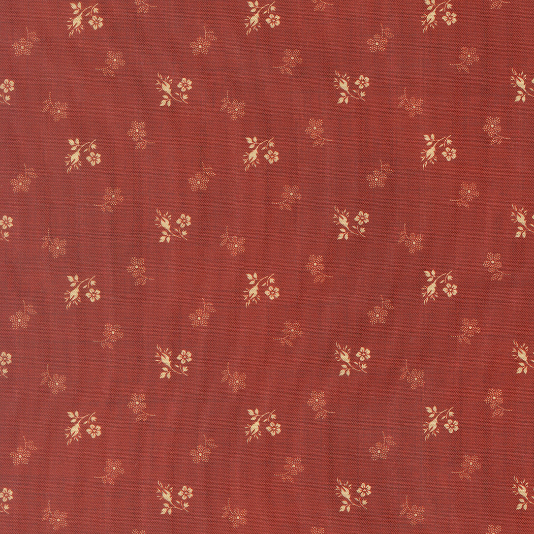 A close-up of a rust red fabric with a repeating pattern of small beige and light brown floral sprigs.