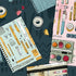 A flat-lay illustration of various stationery items, including notebooks, pens, washi tape, binder clips, and a small paint palette, arranged on a dark teal background with a pink notebook corner visible on the right.