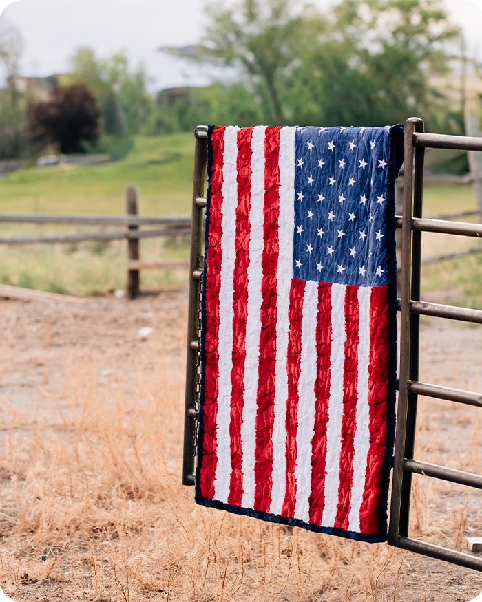 American flag quilt draped over a wooden fence in an outdoor setting with grass and trees.