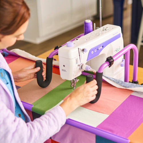 A person using a white and purple sewing machine with purple armrests and black foam grips on a colorful patchwork quilted table in a bright indoor room.