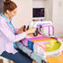 A woman sits at a table using a sewing machine with a colorful fabric panel. The sewing machine is white with purple accents, and the fabric features multiple bright colors including pink, orange, green, blue, and purple.