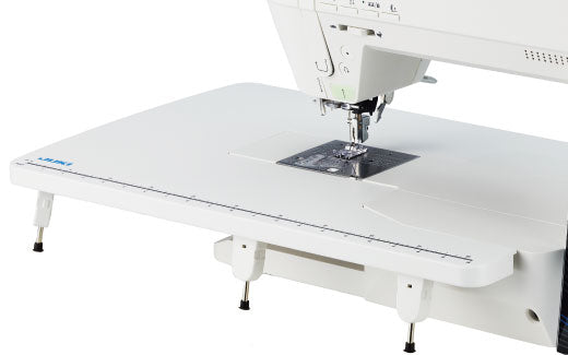 A close-up of a white Juki sewing machine showing the needle area, presser foot, and a large flat bed with measurement markings on a white background.