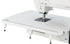 A close-up of a white Juki sewing machine showing the needle area, presser foot, and a large flat bed with measurement markings on a white background.