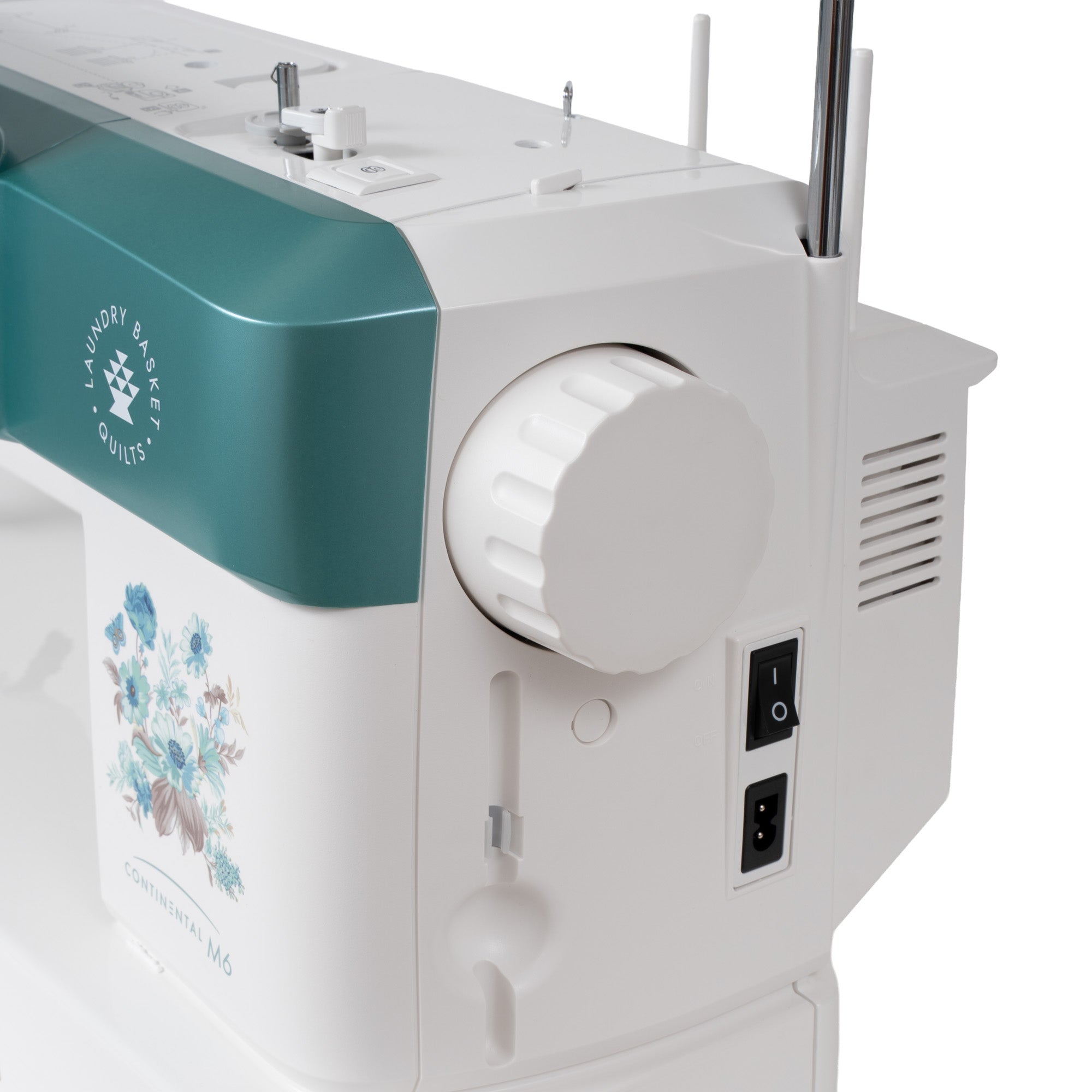A close-up of a sewing machine showing the side panel with a white body, a teal upper section, a large white dial, power switches, and a floral print on the front.