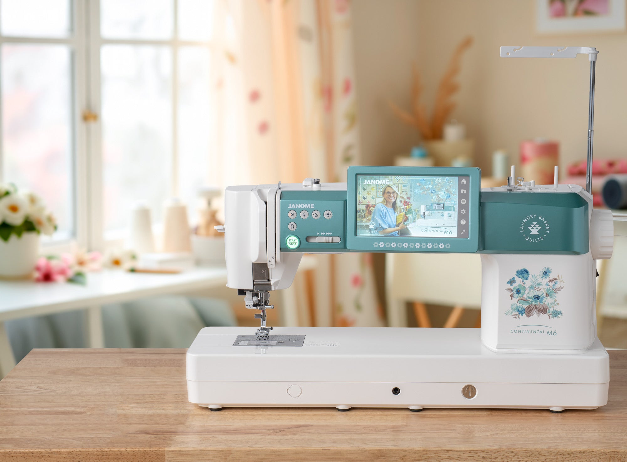 A Janome sewing machine in white and teal sits on a wooden table in a bright, softly blurred craft room setting.