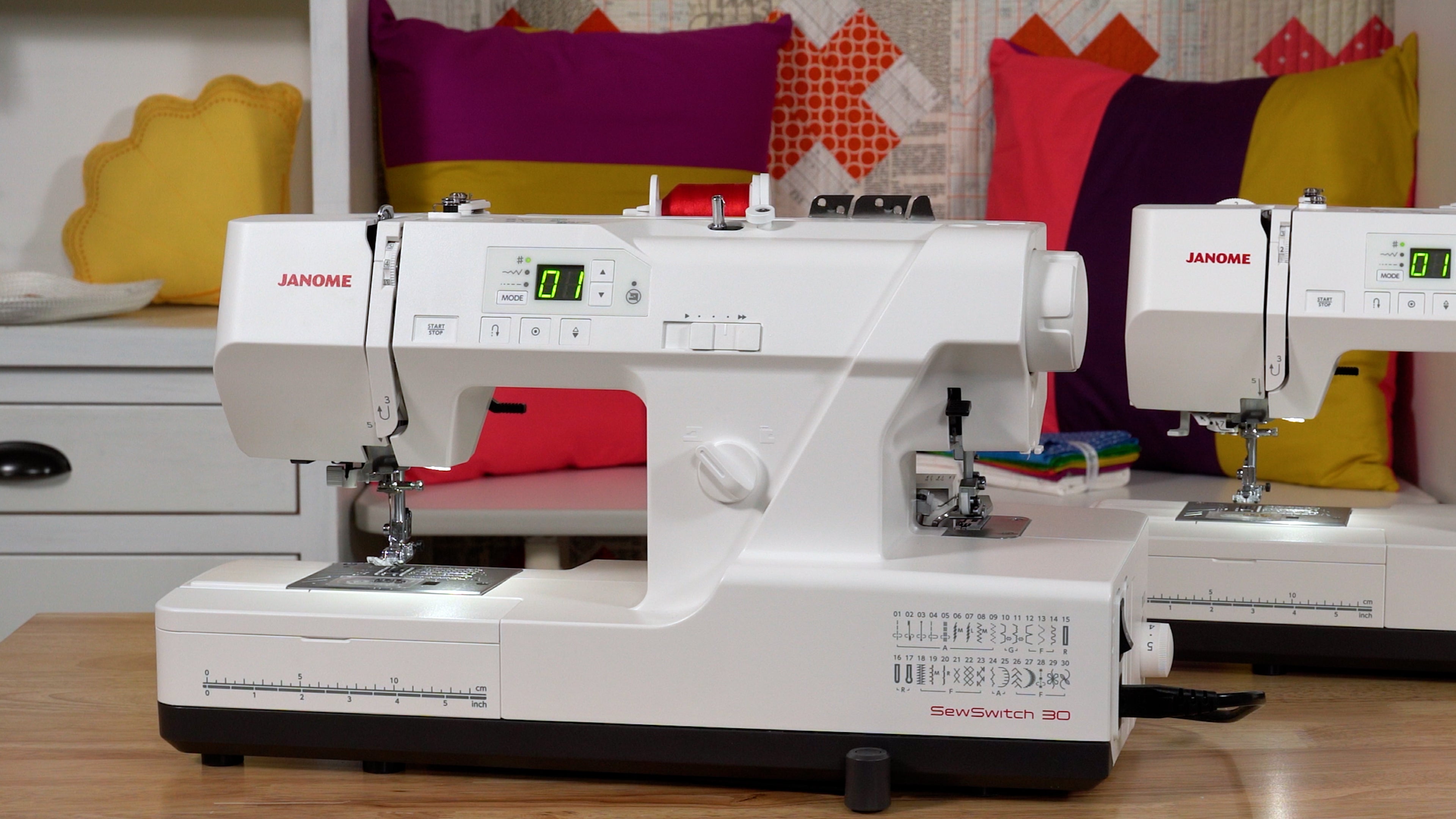 A white Janome SewSwitch 30 sewing machine sits on a wooden table in a sewing room setting, with other sewing machines and colorful quilts visible in the background.
