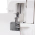 Close-up of a white sewing machine with a metallic presser foot and needle assembly, showing part of the machine body and a white fabric surface.