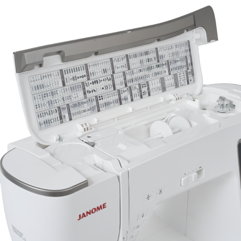 A white Janome sewing machine with its lid open, showing a storage compartment filled with various sewing thread spools and bobbins.