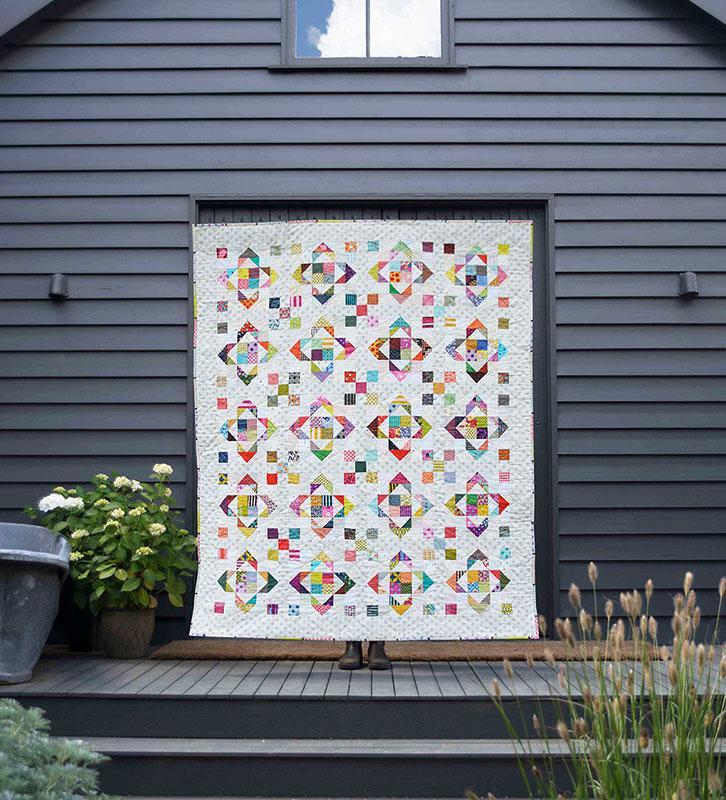 A colorful quilted patchwork blanket or small quilt hanging on a stand on a dark wooden deck in front of a dark gray house exterior.