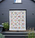 A colorful quilted patchwork blanket or small quilt hanging on a stand on a dark wooden deck in front of a dark gray house exterior.