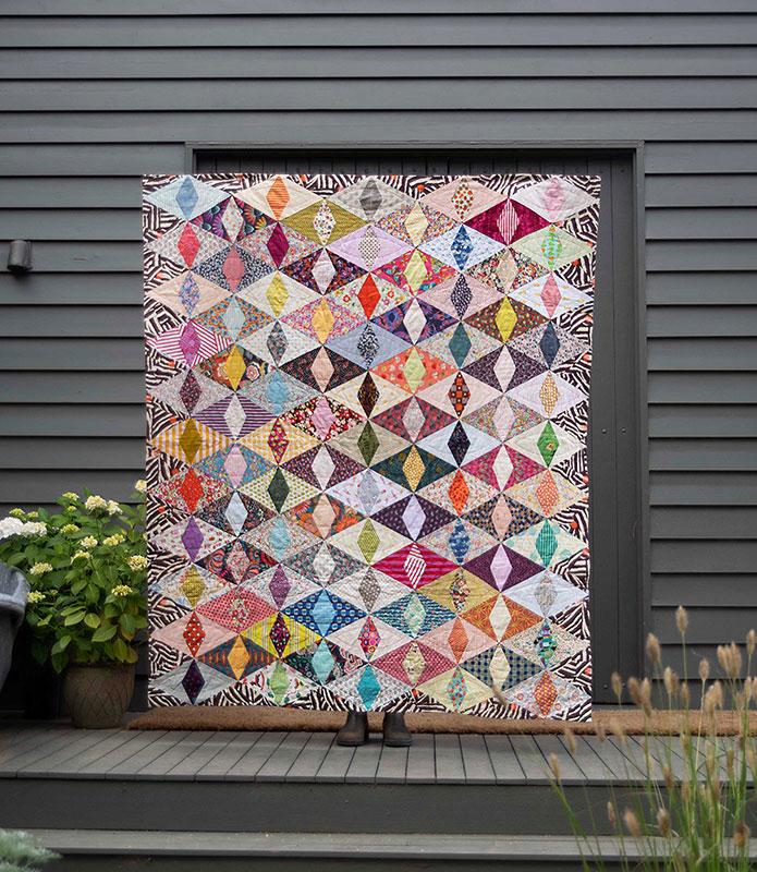 A large patchwork quilt with a colorful geometric diamond pattern is displayed outdoors against a dark gray house siding background on a wooden deck.