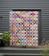 A large patchwork quilt with a colorful geometric diamond pattern is displayed outdoors against a dark gray house siding background on a wooden deck.