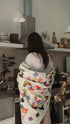 A woman in a kitchen, seen from behind, wrapped in a large, colorful patchwork quilted blanket. The blanket features a white base with various colorful geometric and floral patterns.