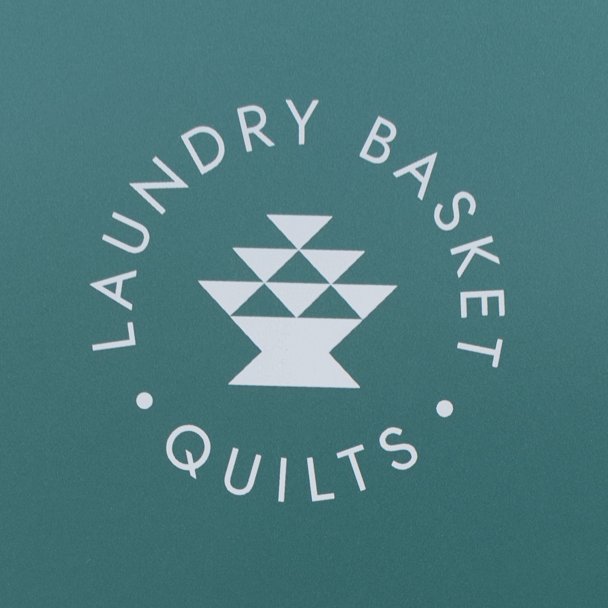 A square teal packaging card or box featuring a white circular logo that reads 'LAUNDRY BASKET QUILTS' around a central geometric basket icon.