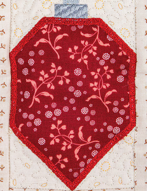 A red fabric ornament-shaped patch with a pink floral and berry pattern, featuring a blue-gray fabric tab at the top, displayed on a cream quilted fabric background.