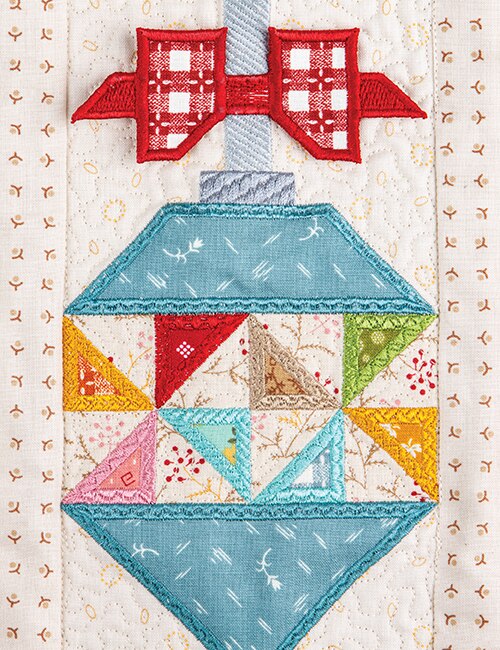 A close-up of a quilted ornament design featuring a red bow at the top, a light blue upper section, and a multicolored geometric patterned lower section on a cream quilted background.
