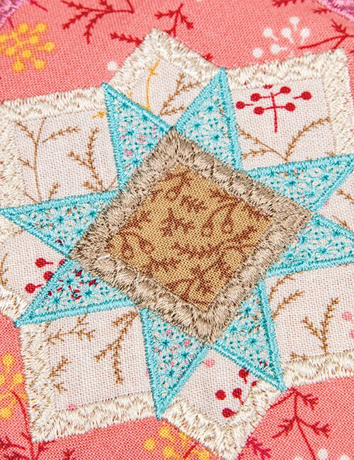 Close-up of a handmade embroidered patch featuring a starburst design with turquoise blue triangles, a tan center square, and cream floral sections on a pink patterned background.