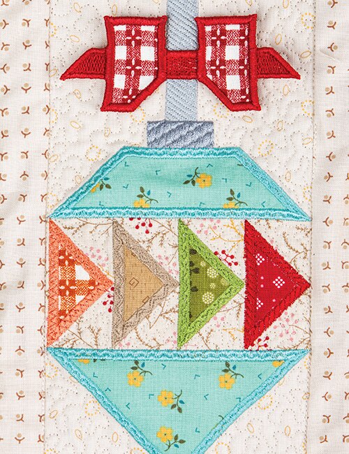 A close-up of a handcrafted quilted ornament applique featuring a red bow, light blue sections with yellow floral prints, and geometric triangle panels in orange, tan, and green on a cream patterned fabric background.