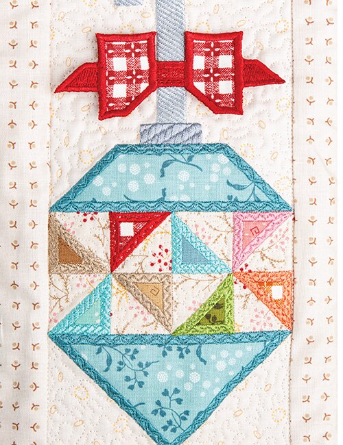 A close-up of a quilted fabric ornament design with a red bow at the top, a light blue diamond-shaped body, and multicolored triangular patchwork sections including pink, green, orange, beige, and brown.