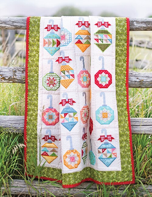 A quilted blanket with a colorful patchwork ornament design is draped over a rustic wooden fence in a grassy outdoor setting.