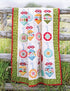 A quilted blanket with a colorful patchwork ornament design is draped over a rustic wooden fence in a grassy outdoor setting.