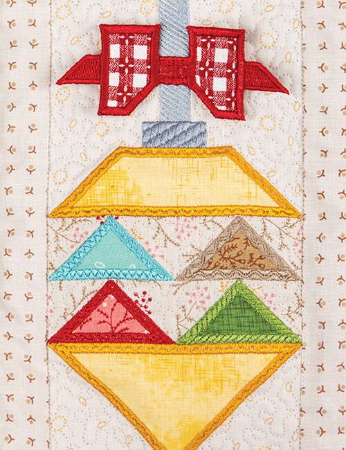 A close-up of a quilted fabric ornament design with a red bow at the top, a yellow diamond shape, and smaller geometric sections in blue, green, and brown on a cream patterned fabric background.