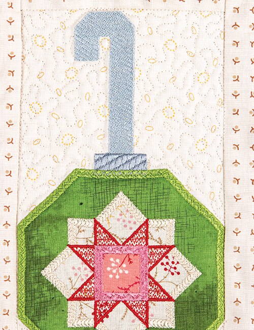 A close-up of a quilted textile featuring a green octagonal shape with a pink and red star motif, a pale blue J-shaped element above it, and an off-white quilted background with embroidered details.