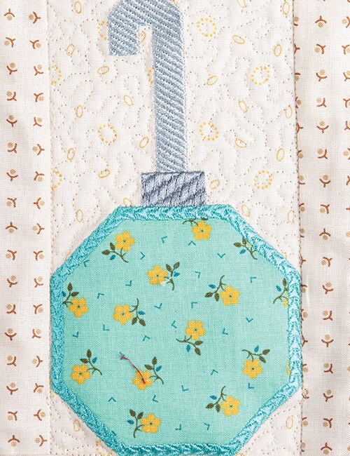 A close-up of a quilted fabric ornament applique featuring a mint green octagonal body with yellow floral print and a light blue stitched handle, set against a cream quilted background with small brown floral motifs.